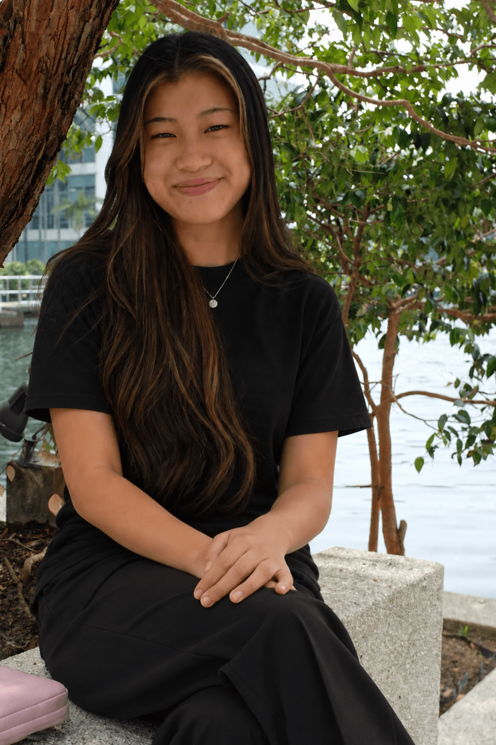 Smiling young woman with long brown hair sits on a stone bench near water.