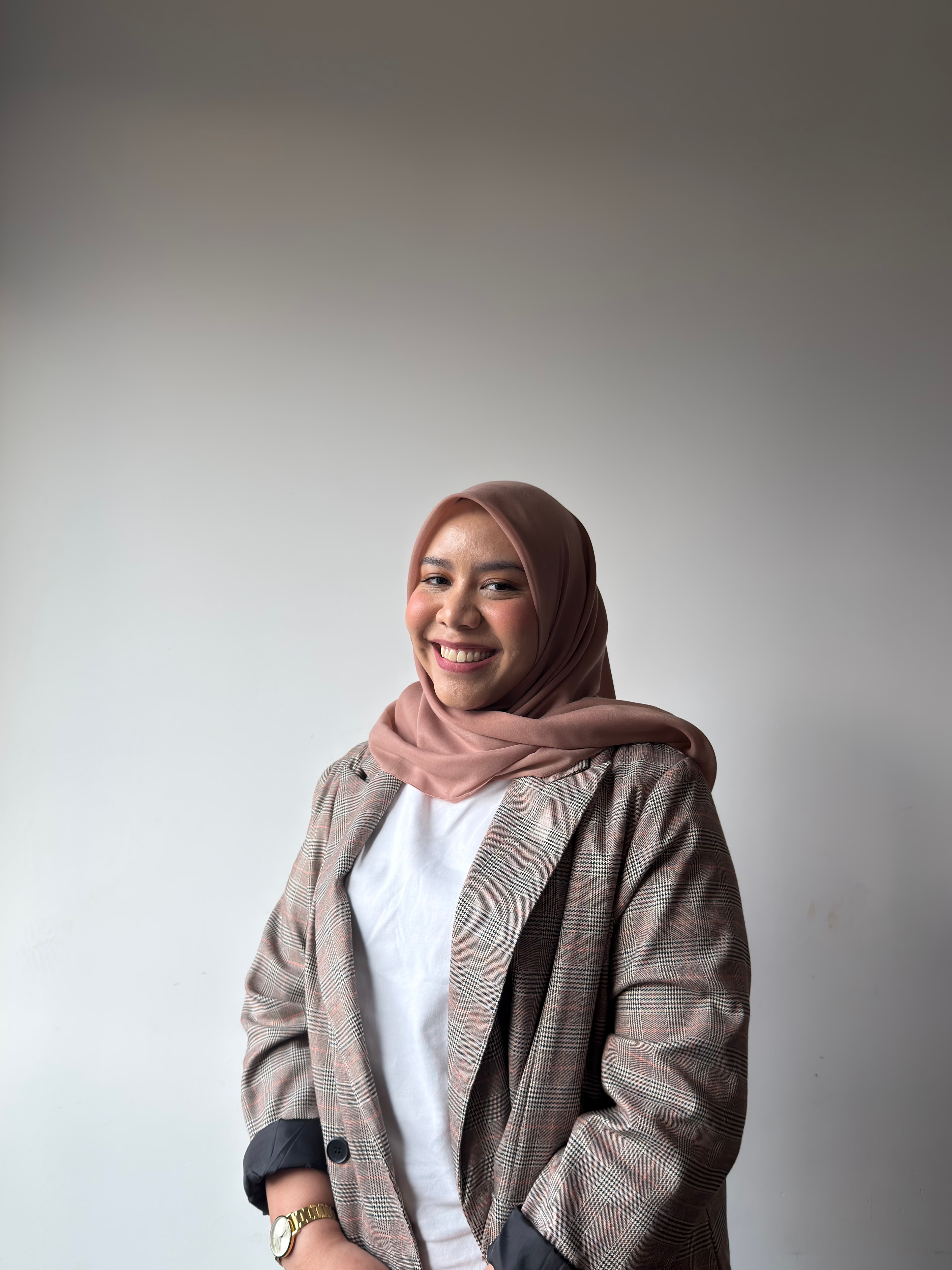 Smiling woman wearing a pink hijab and plaid blazer against a plain white wall.