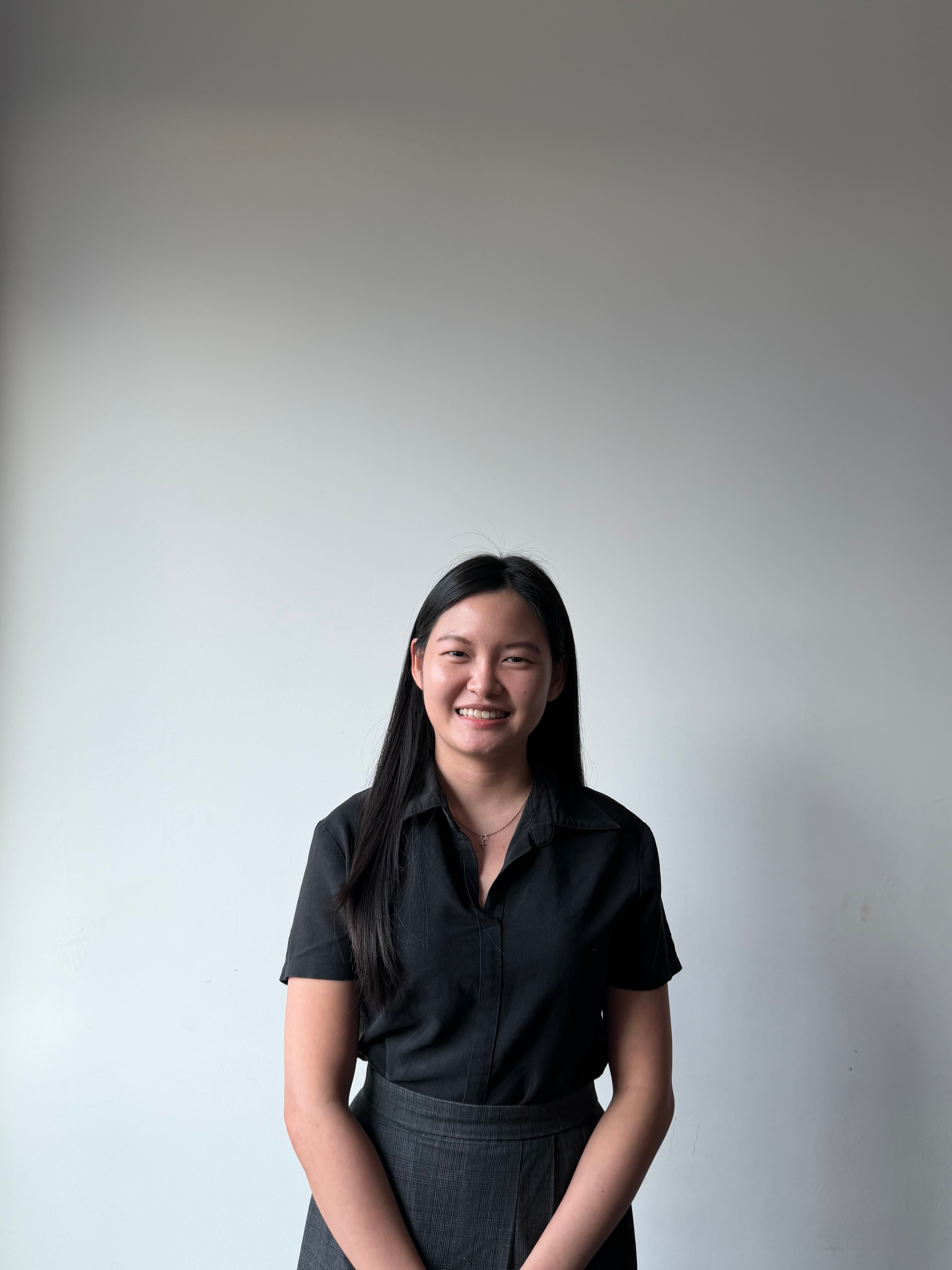 Smiling young woman with long black hair wearing a black shirt and grey plaid skirt.