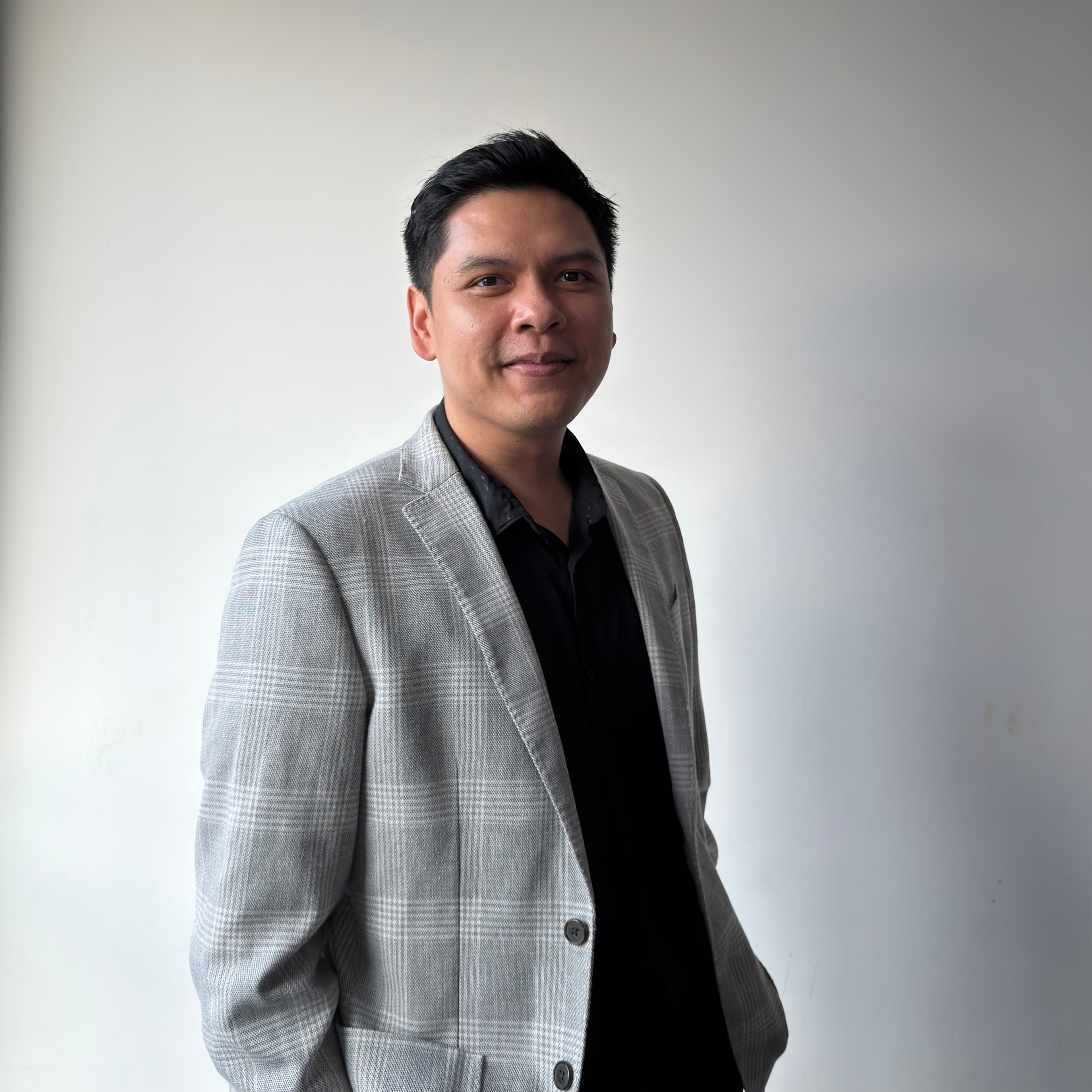 A smiling man wearing a grey plaid blazer and black shirt against a white background.