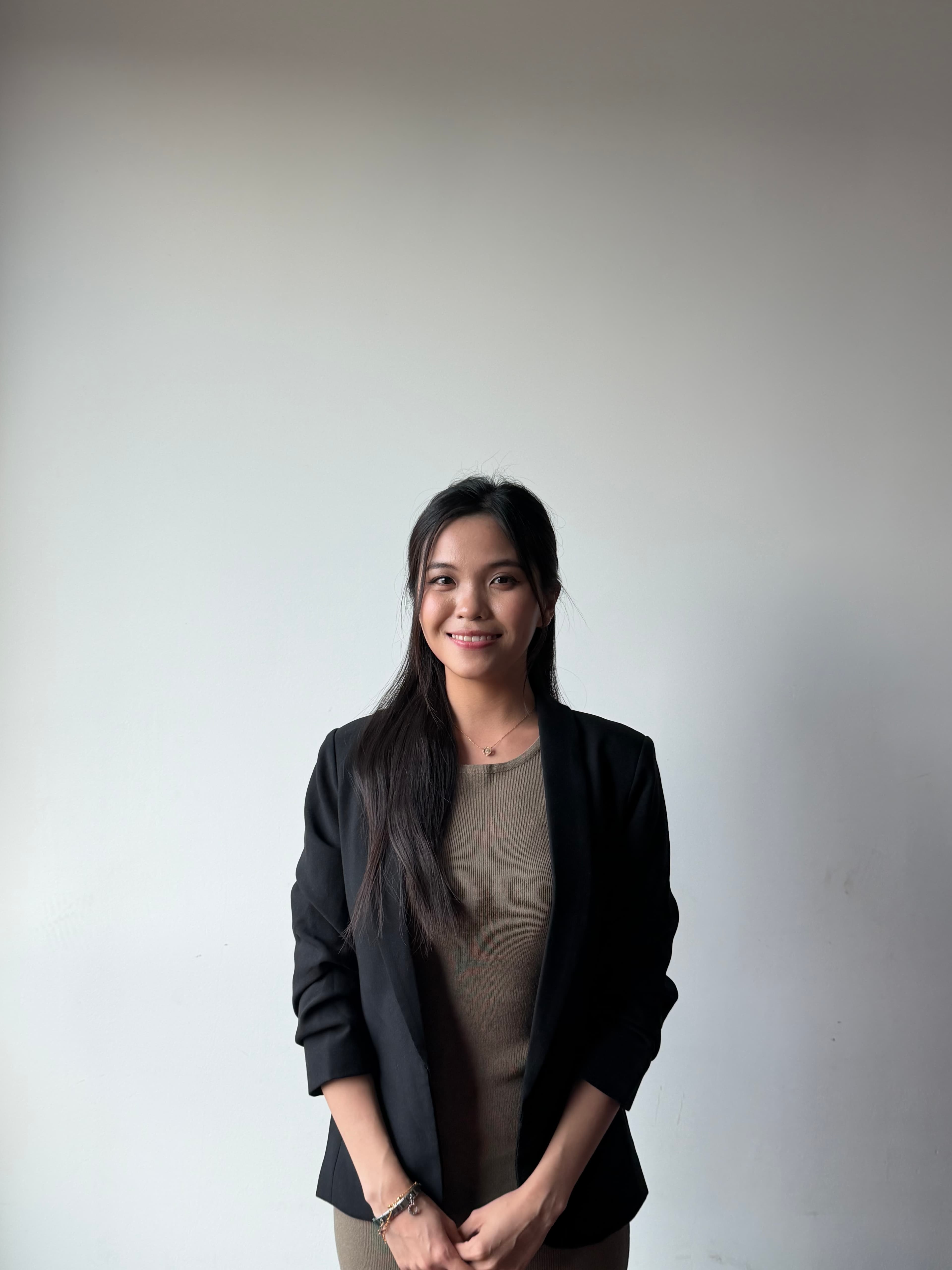 Smiling woman with long dark hair wearing a black blazer against a white wall.